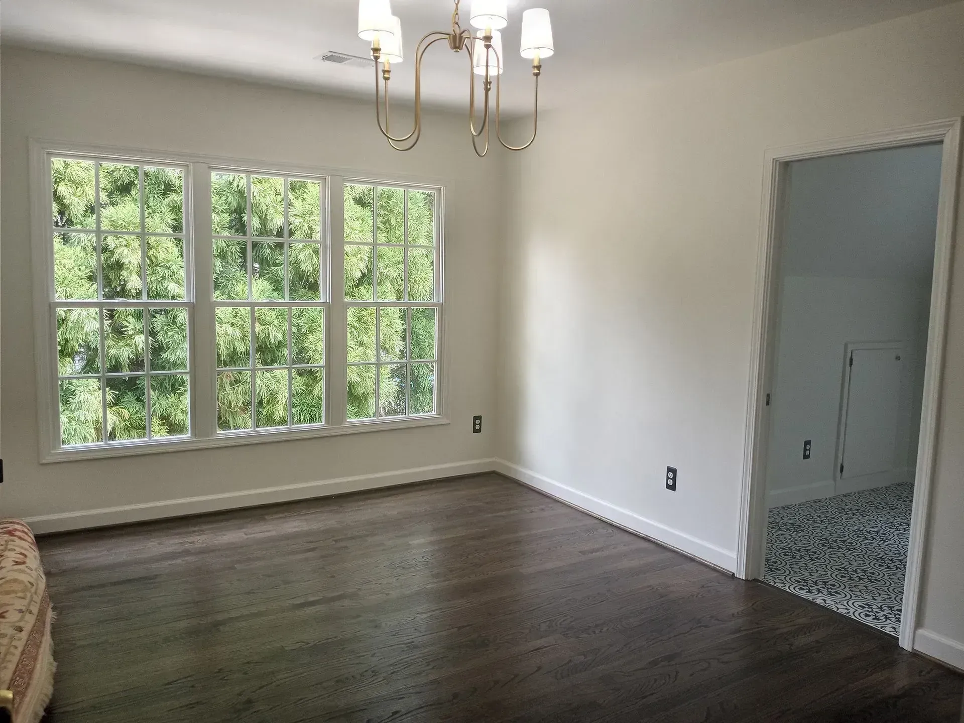 Empty bedroom with dark wood floors, white walls, large window, and an open doorway to a tiled bathroom.