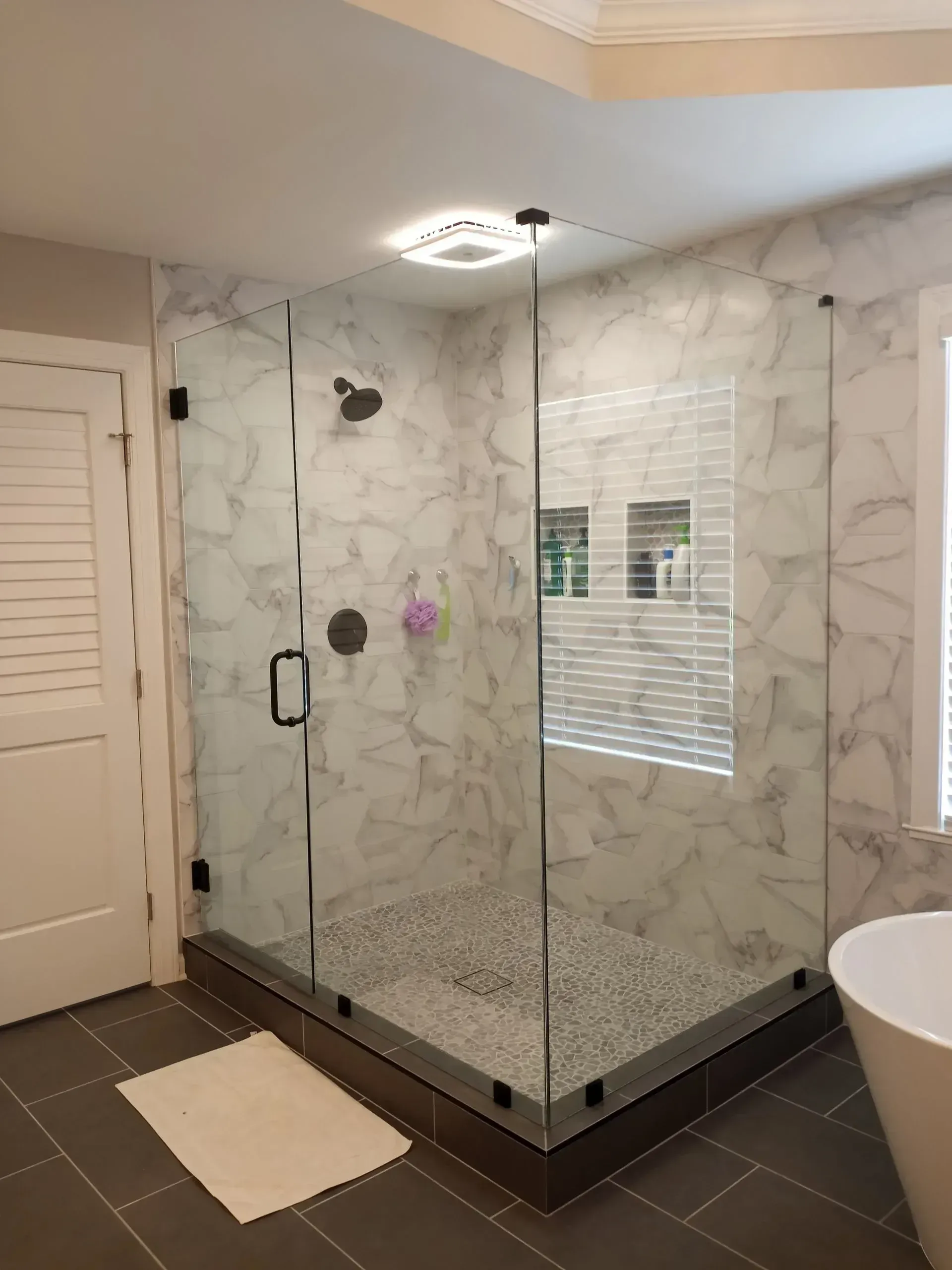 Marble-tiled bathroom with glass shower enclosure, dark floor tiles, and a window with blinds