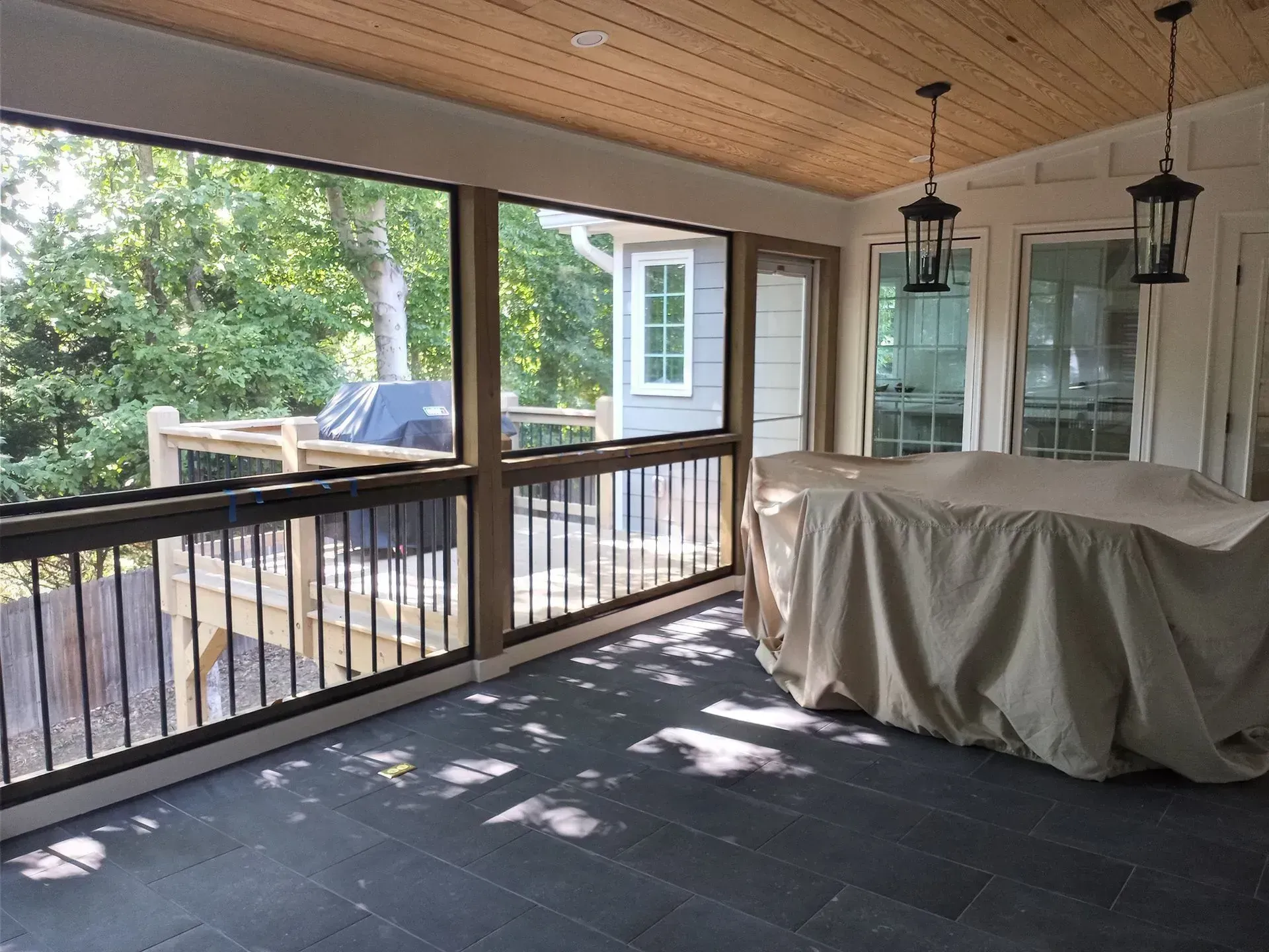 Covered porch with black railing, hanging lanterns, and a table draped in a gray cover overlooking a yard