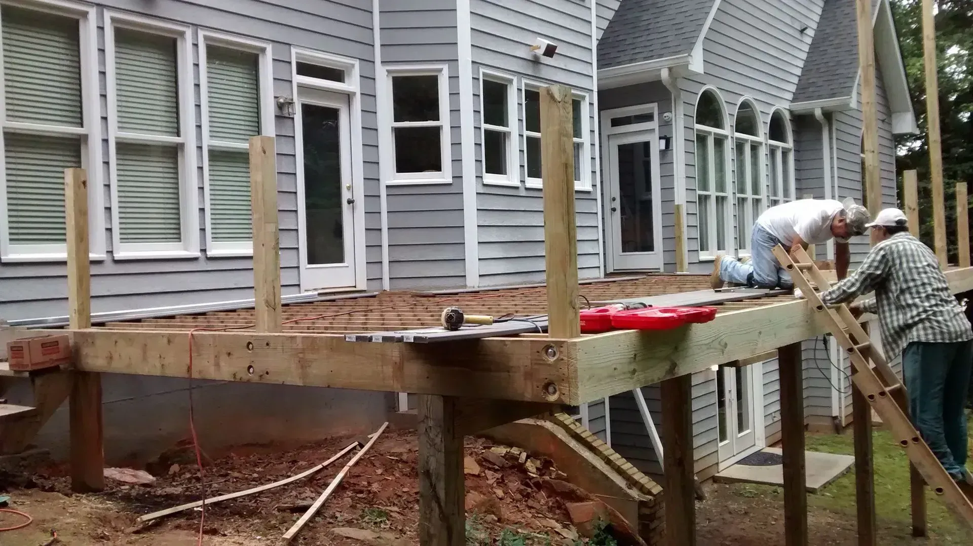 Two workers building a raised wooden deck beside a gray house.