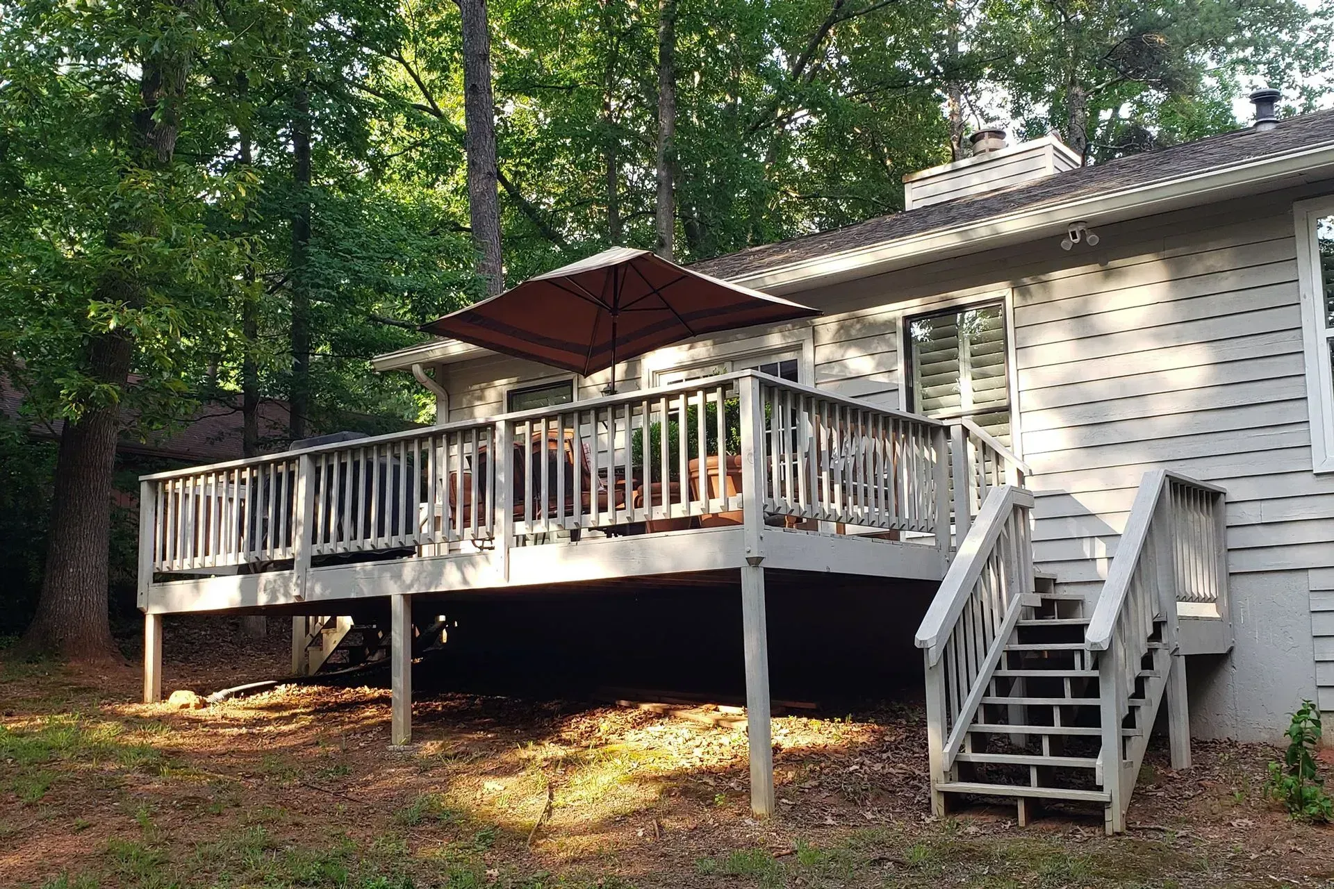 White house deck with stairs, railing, and red patio umbrella in a wooded yard.