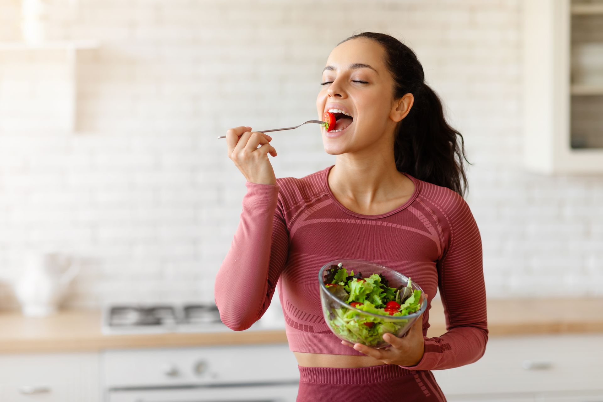 Woman in a pink top tasting salad in a bright kitchen, holding a bowl of greens.