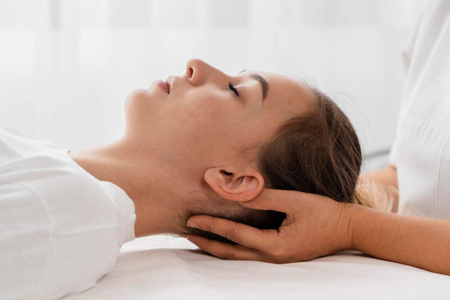 Person receiving neck massage. Therapist's hands support head, patient eyes closed, white setting.