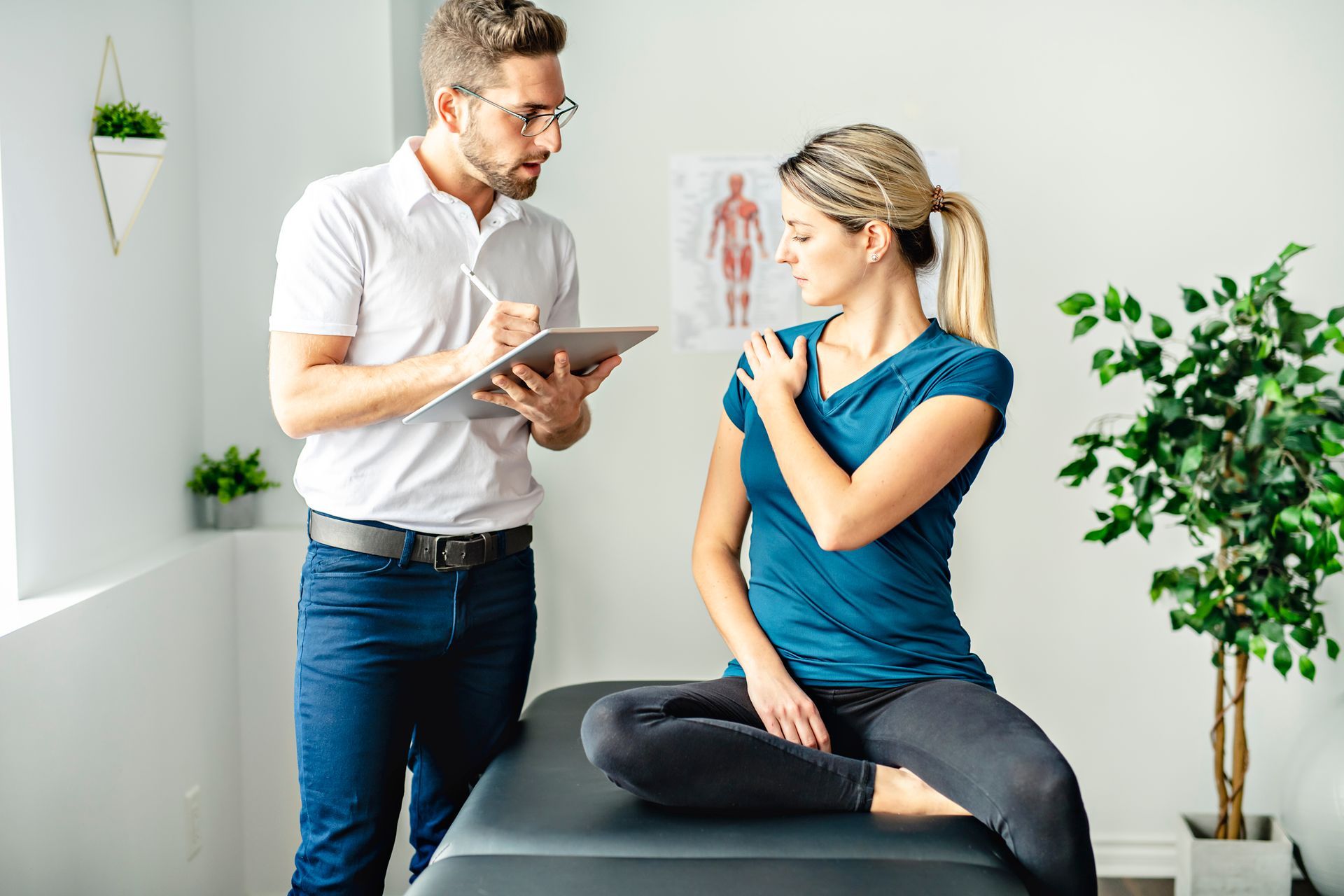 A medical professional examines a patient's shoulder in a clinic; the patient is seated on an examination table.