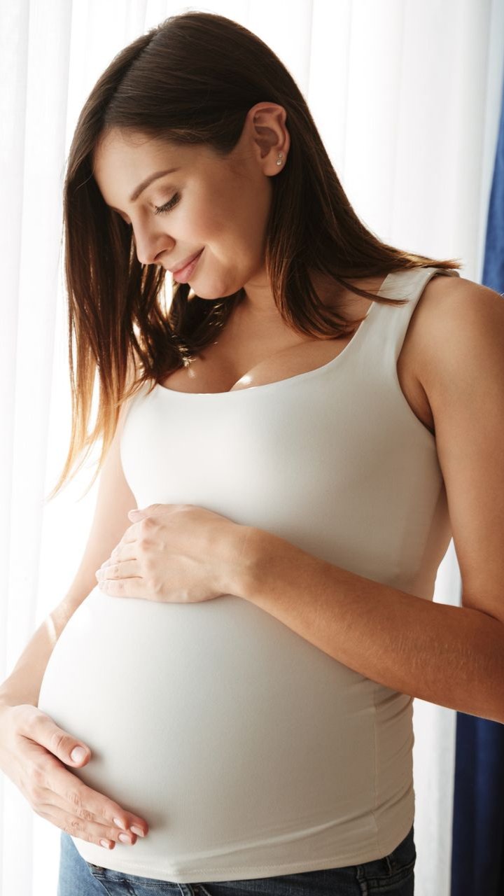 Pregnant person in a white tank top gently holding their belly by a bright window