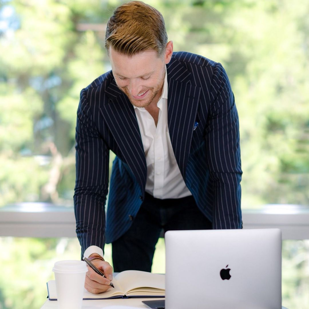 Man in blue striped blazer writes in notebook at a desk with laptop and coffee, window in background.