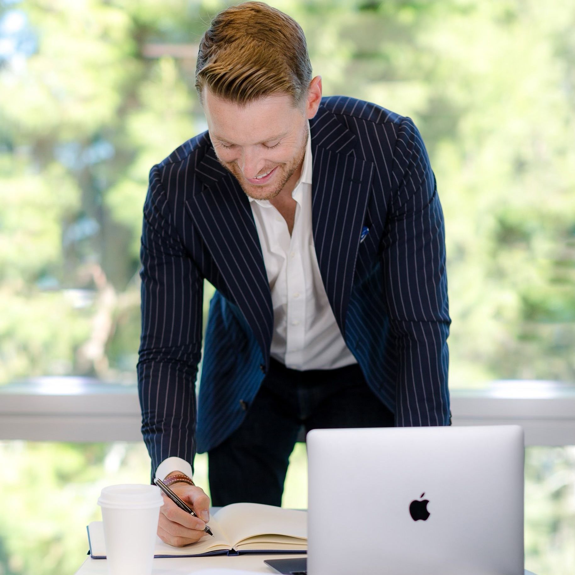 Man in blue striped blazer writes in notebook at a desk with laptop and coffee, window in background.