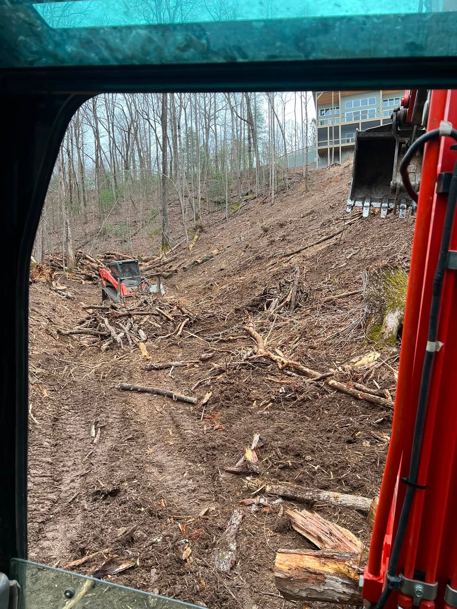 View from a red excavator cab over a muddy wooded slope with fallen branches and a shovel in the distance