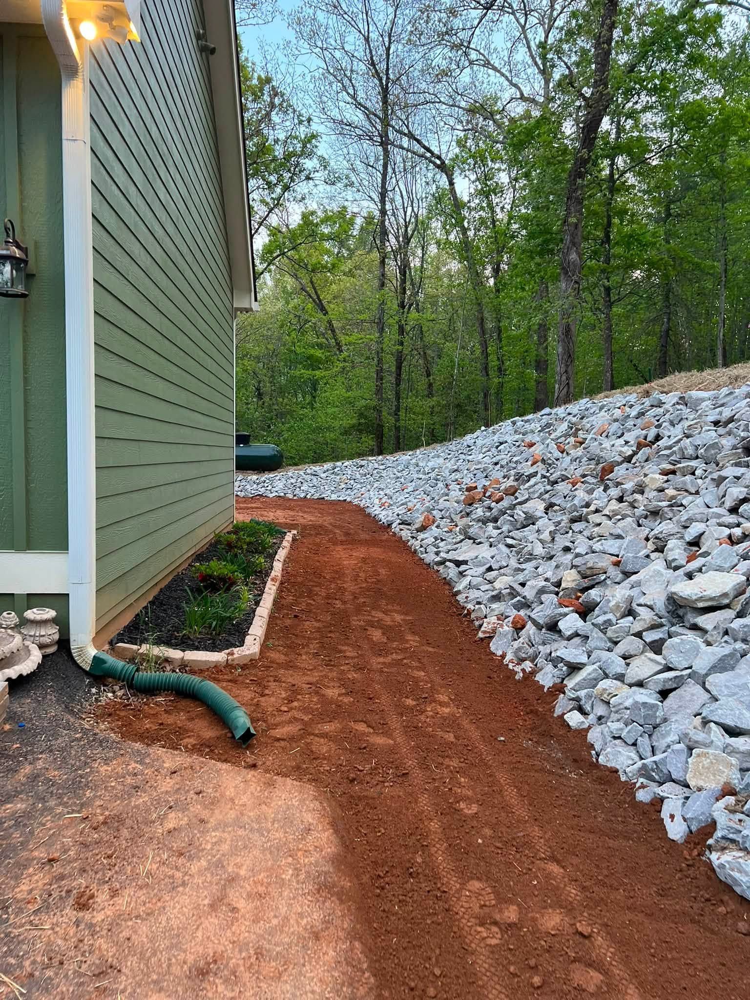 Side yard with green house wall, red mulch path, and a sloped pile of gray rocks beside trees.