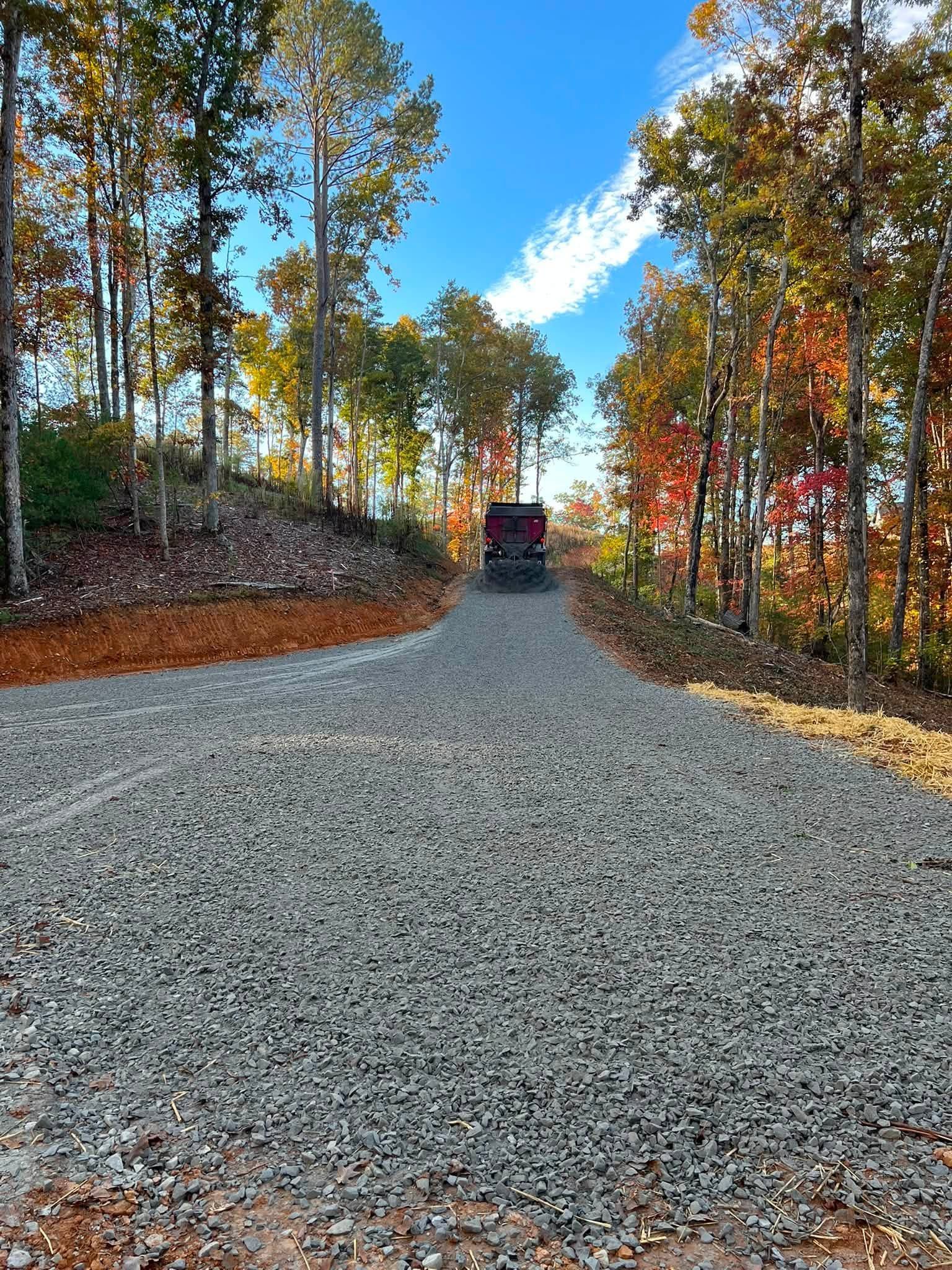 Gravel road curving uphill through autumn trees under a blue sky, with a vehicle in the distance