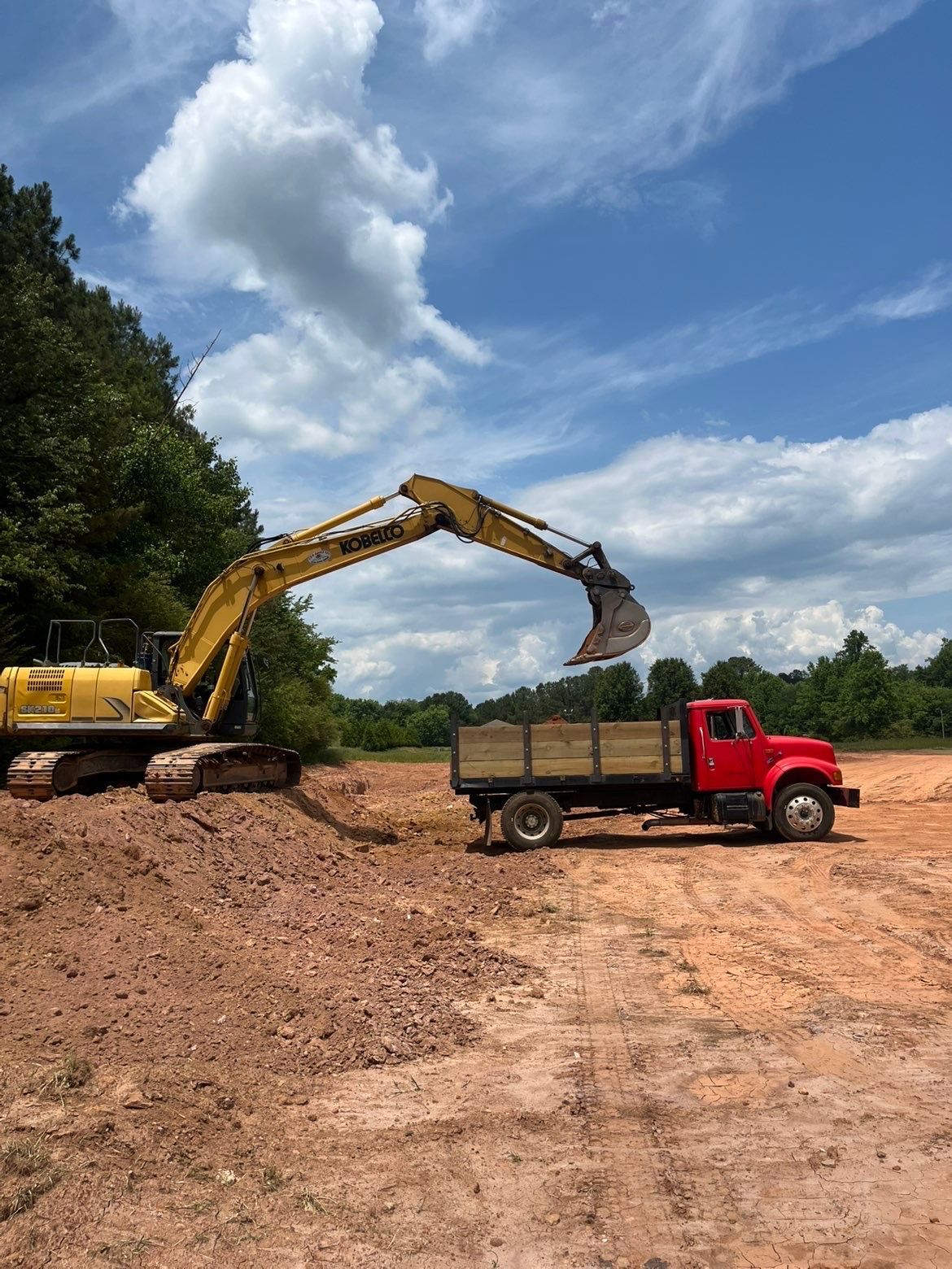 Yellow excavator loading dirt into a red dump truck at a construction site under a blue sky