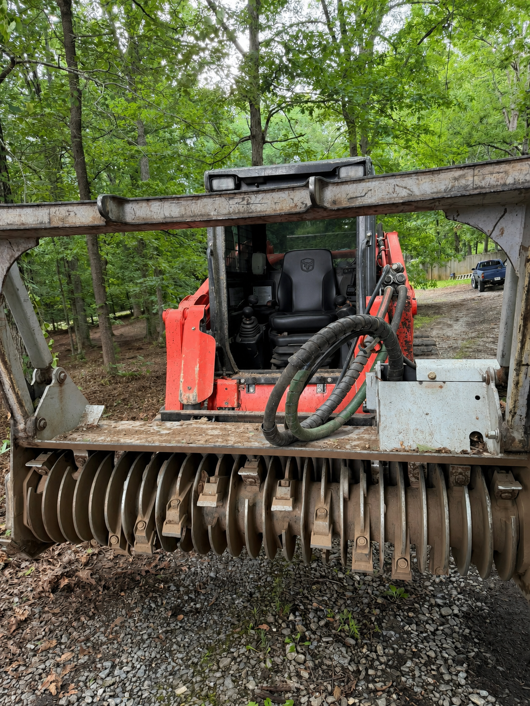 Orange skid steer with brush cutter attachment in a wooded area, clearing gravel and leaves