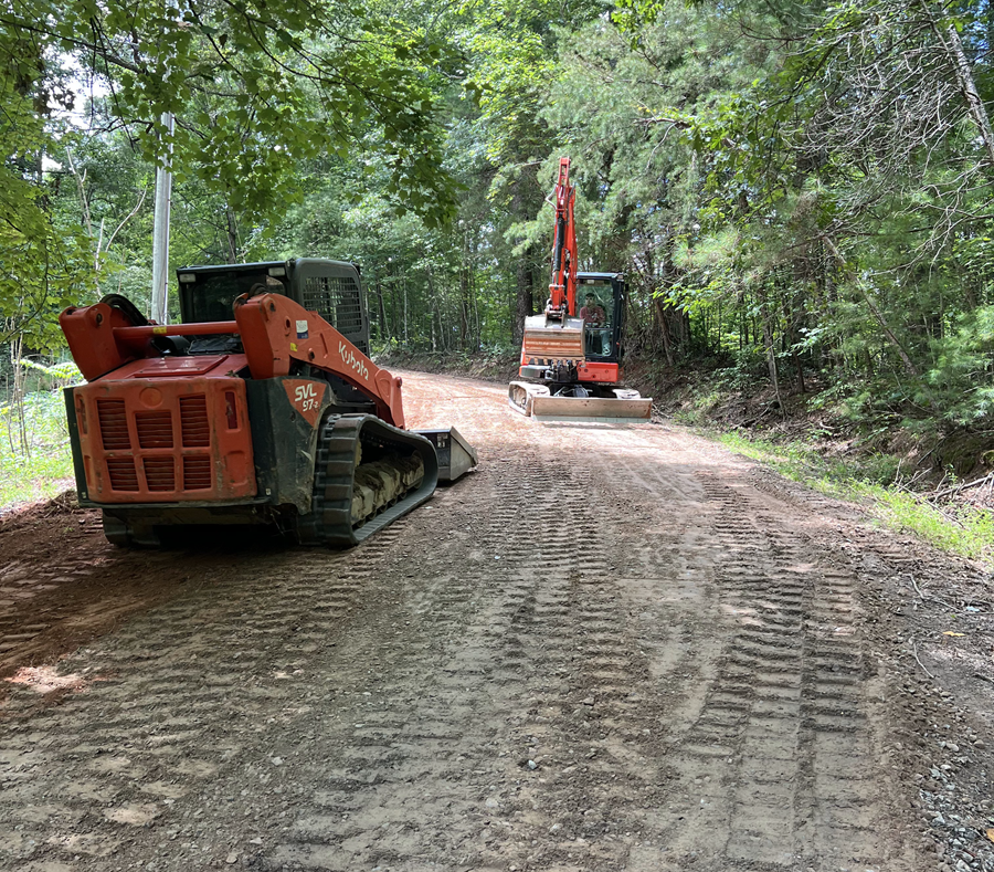 Two orange excavators on a dirt road in a wooded area, one in the foreground and one farther ahead.