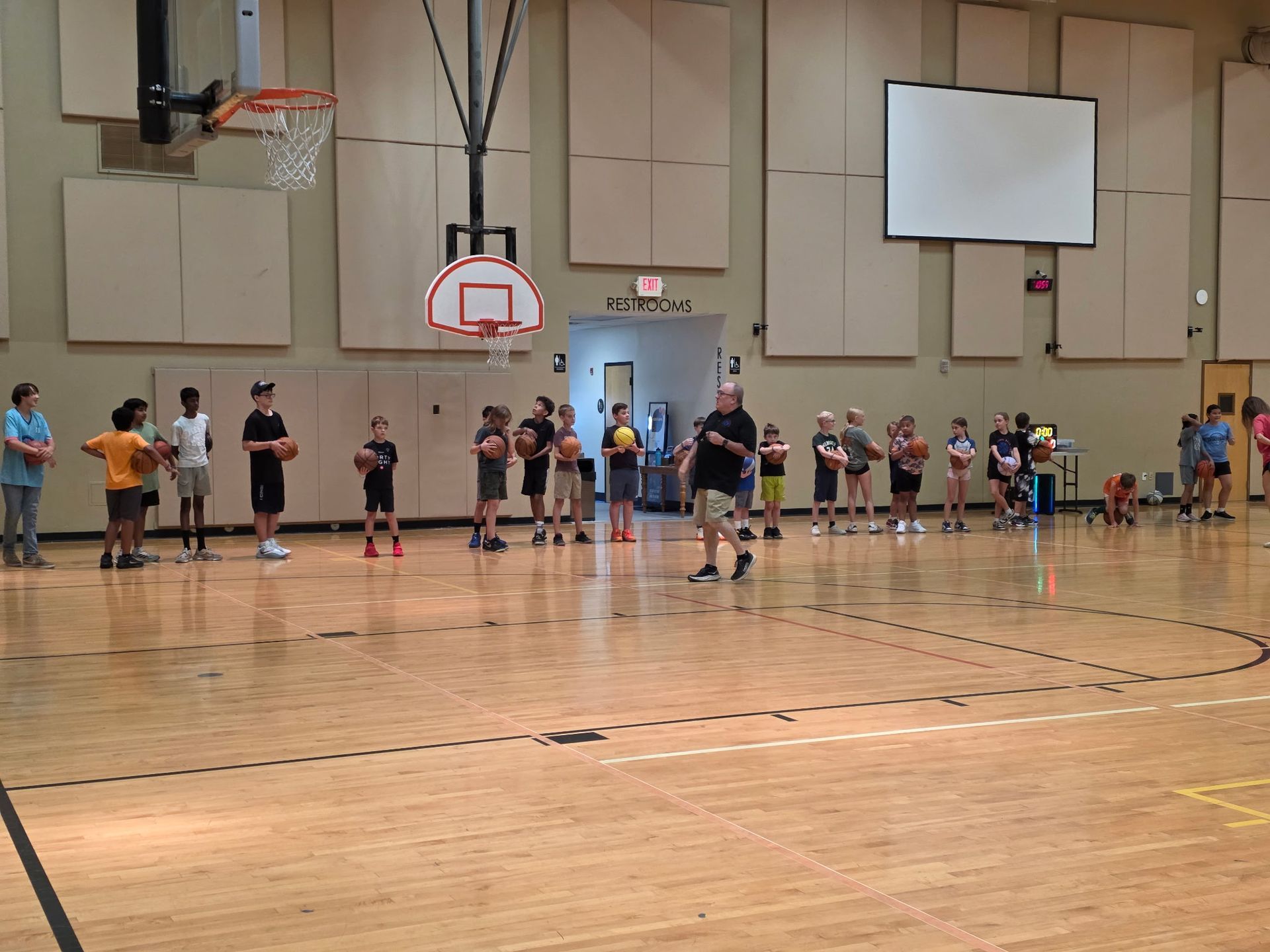 Basketball gym with players lined up near the court and hoop during practice.