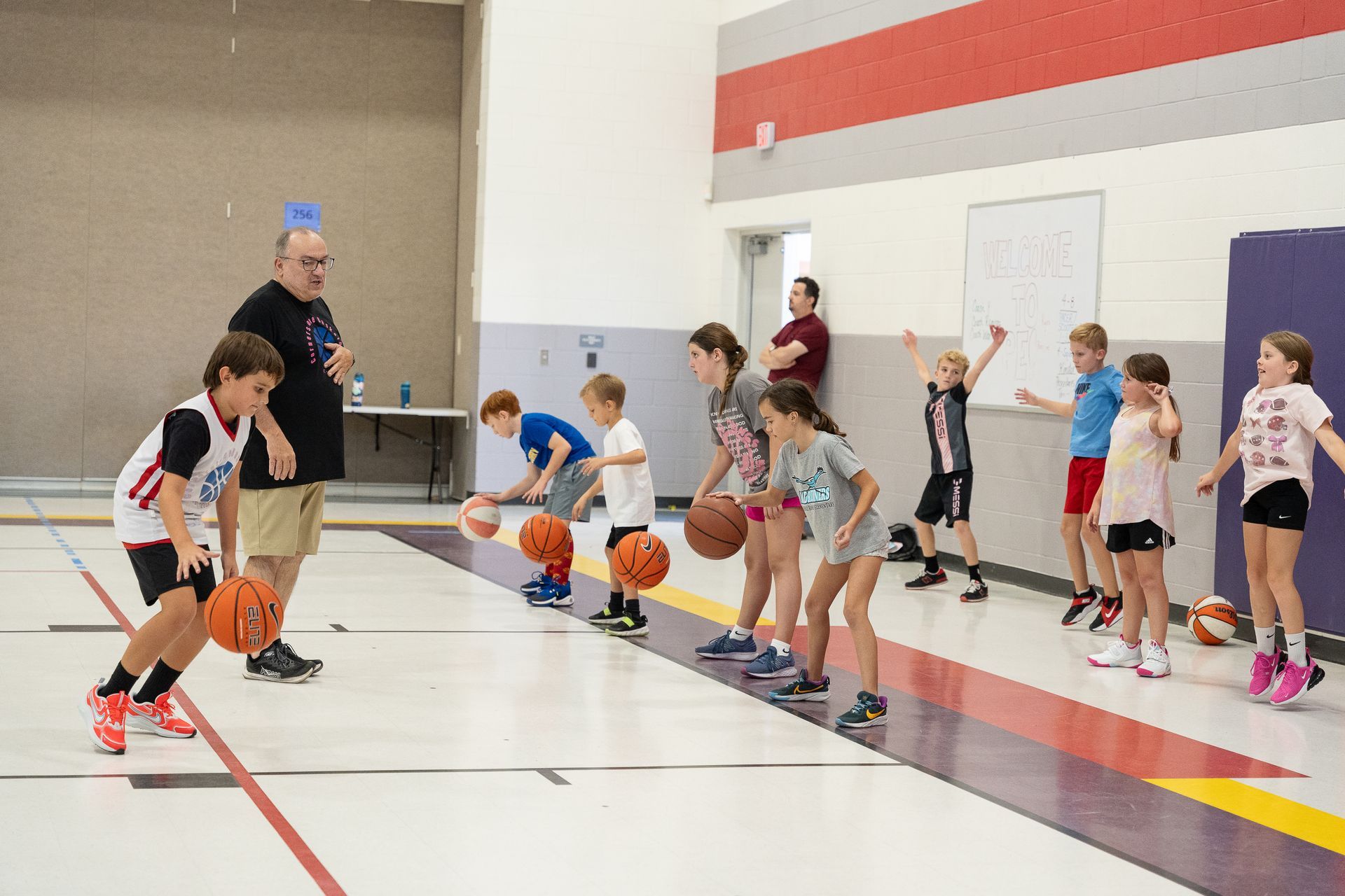 Children dribbling basketballs during a gym practice with a coach watching nearby