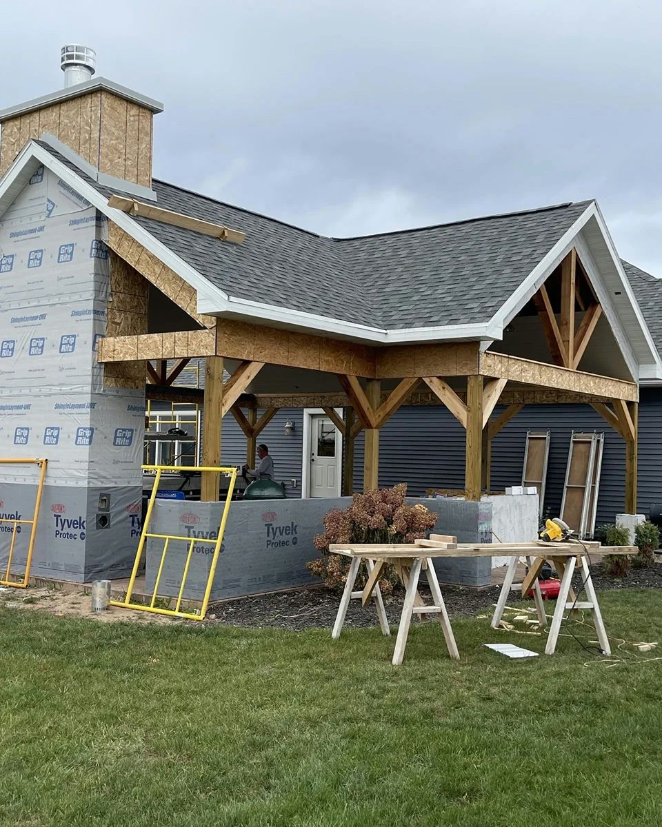 Construction of a wooden pergola attached to a house with gray siding. Work tools visible, overcast sky.