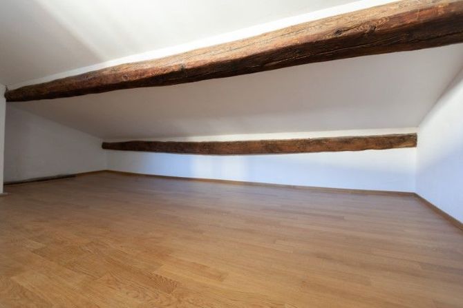Empty attic room with sloped white walls, exposed wooden beams, and a hardwood floor