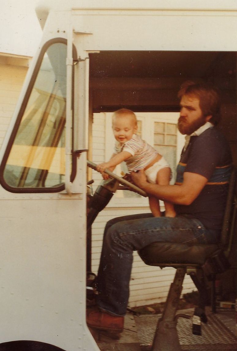 Man holding a baby at the steering wheel of a white truck. Both are smiling, inside the truck's cabin.