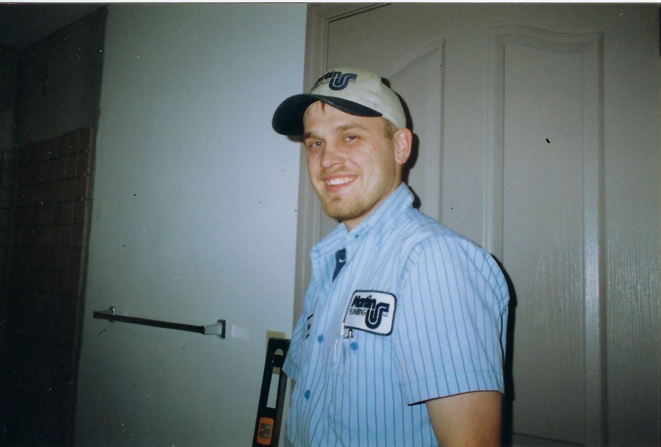 Man in work uniform and cap smiles. Standing near towel rack, door, and a level.