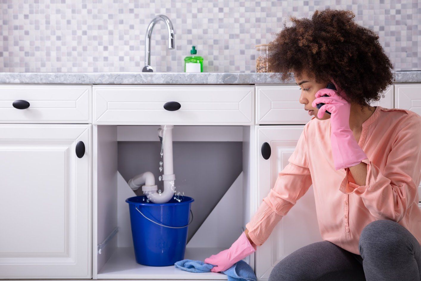 A woman is talking on a cell phone while cleaning under a sink.