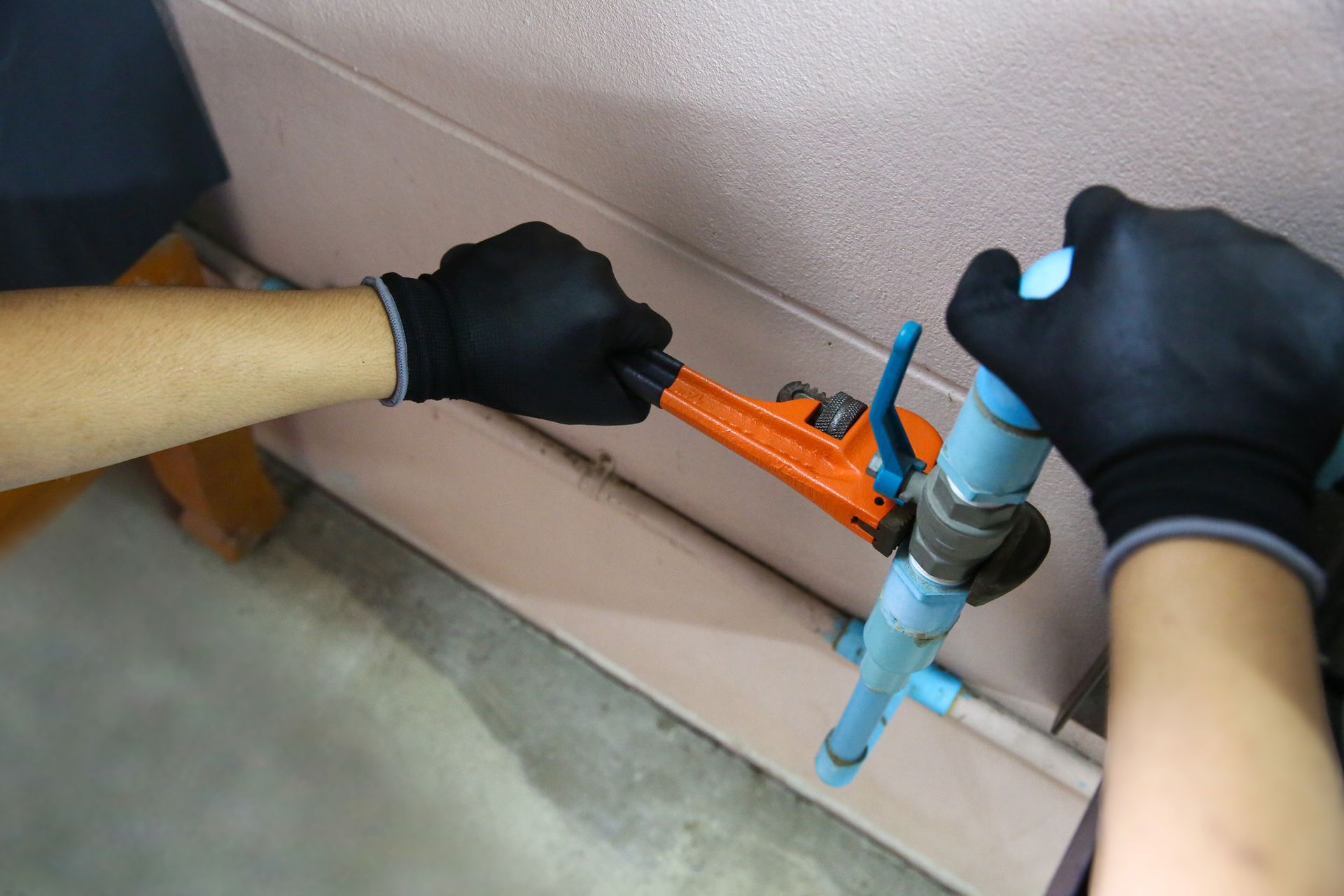 Gloved hands using an orange pipe tool on blue plumbing against a wall with a gray floor