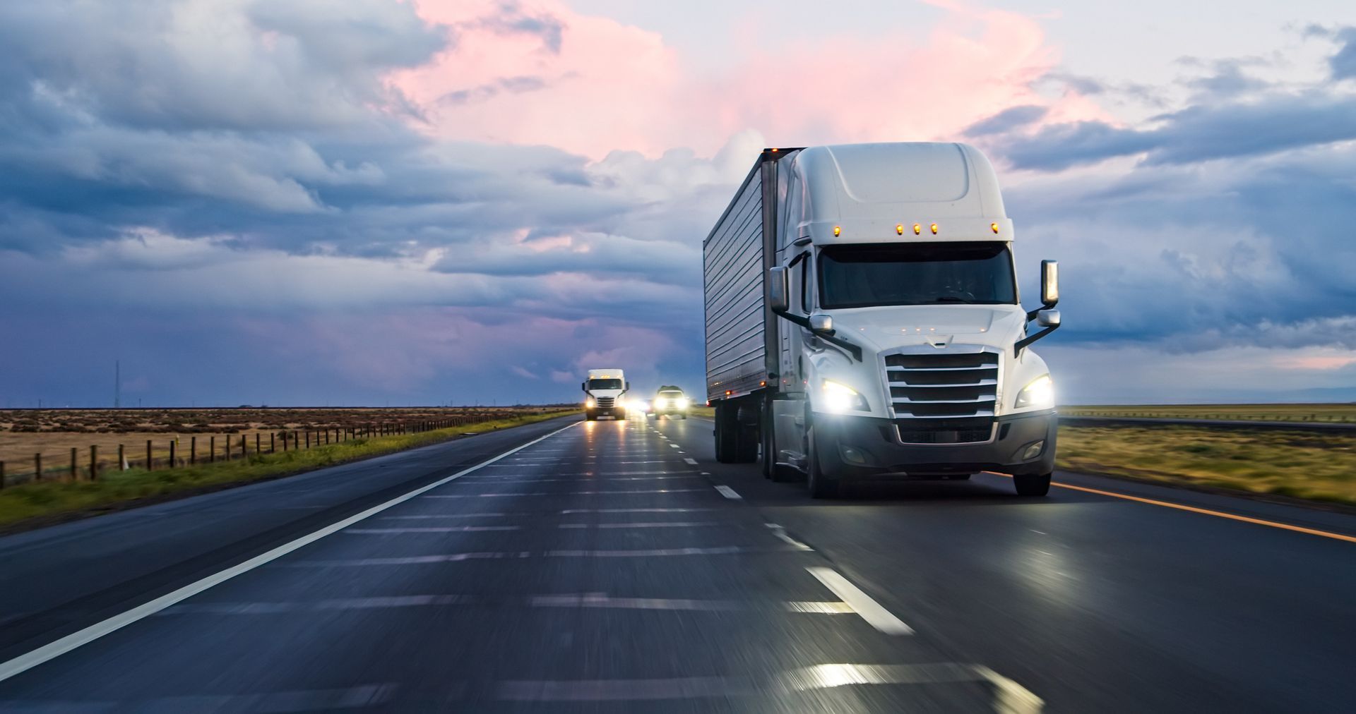 Two semi-trucks driving on a wet highway under a cloudy sky at dusk. Two semi-trucks driving on a wet highway under a cloudy sky at dusk.
