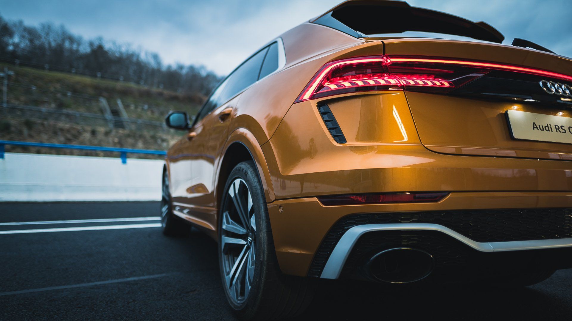 Gold Audi RS Q8 SUV parked on a road, rear view. Tail lights illuminated, exhaust visible.