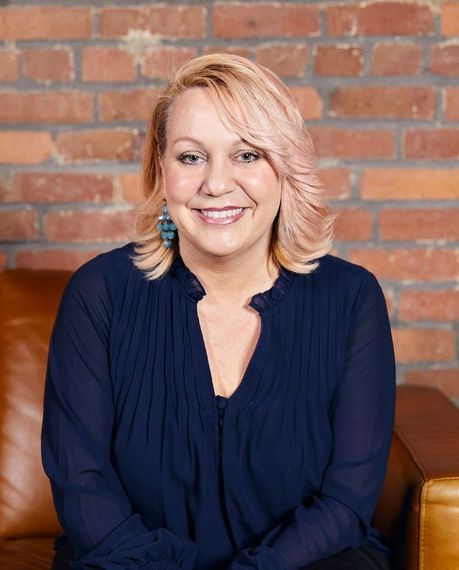 A woman is smiling while sitting on a couch in front of a brick wall.
