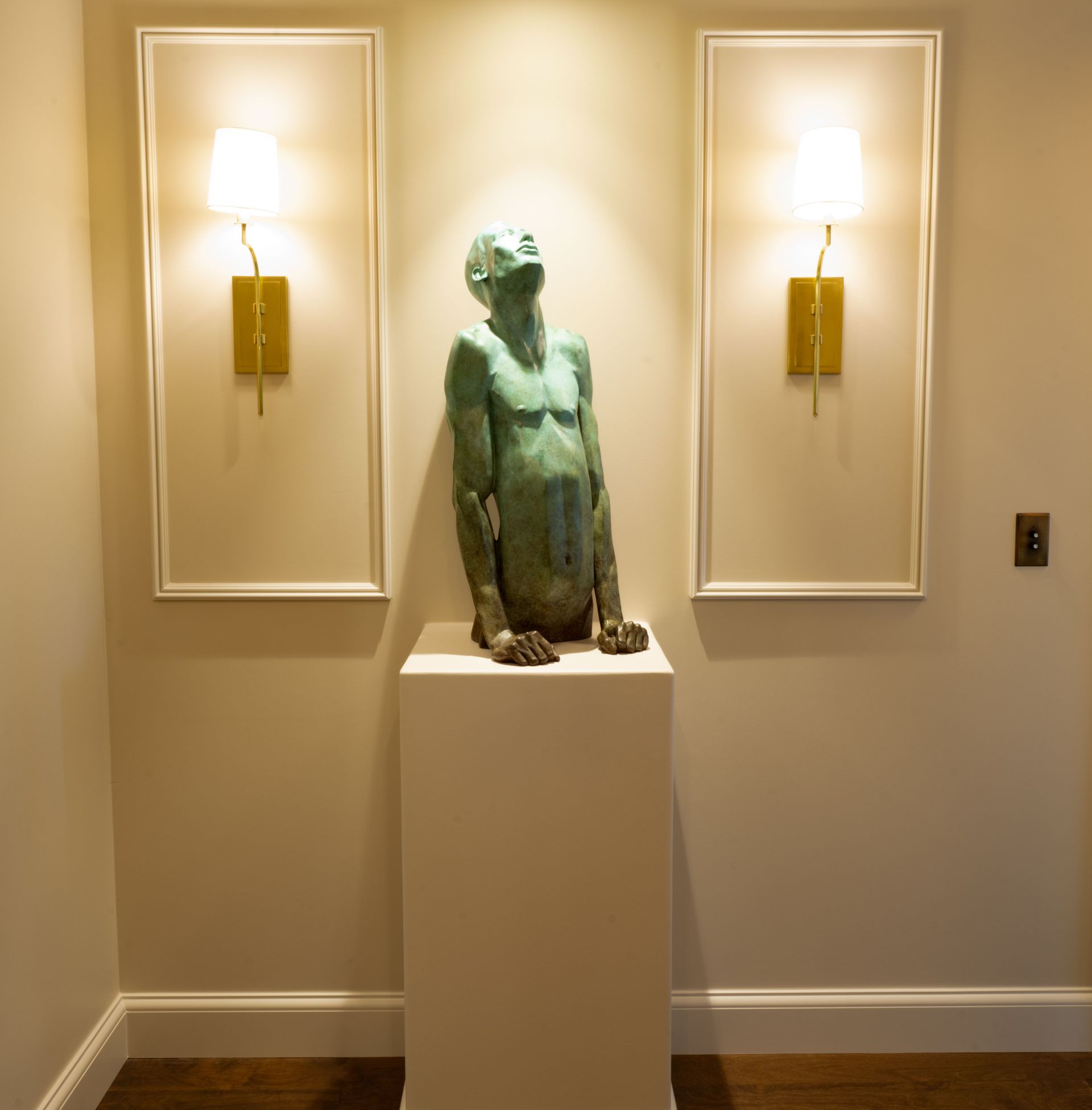 Bronze sculpture on pedestal between two wall sconces. Beige walls, light brown trim.