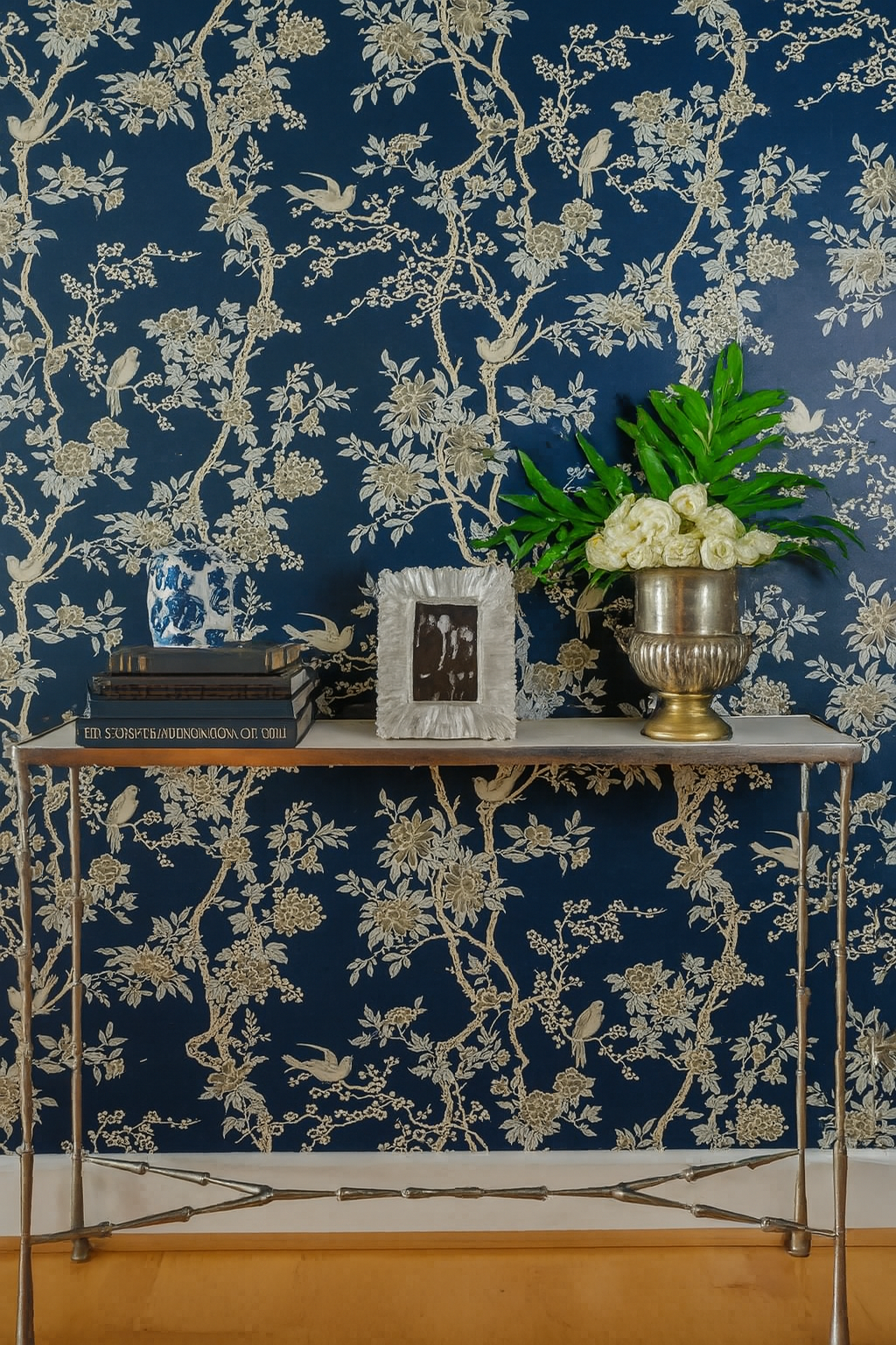 Console table with books, vase of flowers, and a framed photo against a blue floral wallpaper backdrop.