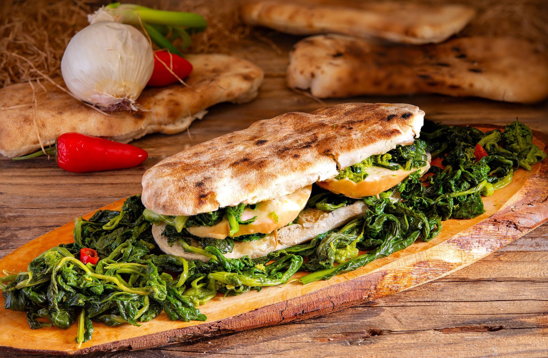 Panino grigliato con verdure e formaggio su pane rustico, con aglio e peperoni rossi sullo sfondo.
