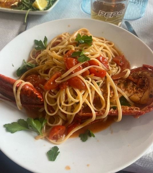 Spaghetti con astice e salsa di pomodoro su un piatto bianco, con guarnizione di prezzemolo e una bottiglia di Pepsi sullo sfondo.