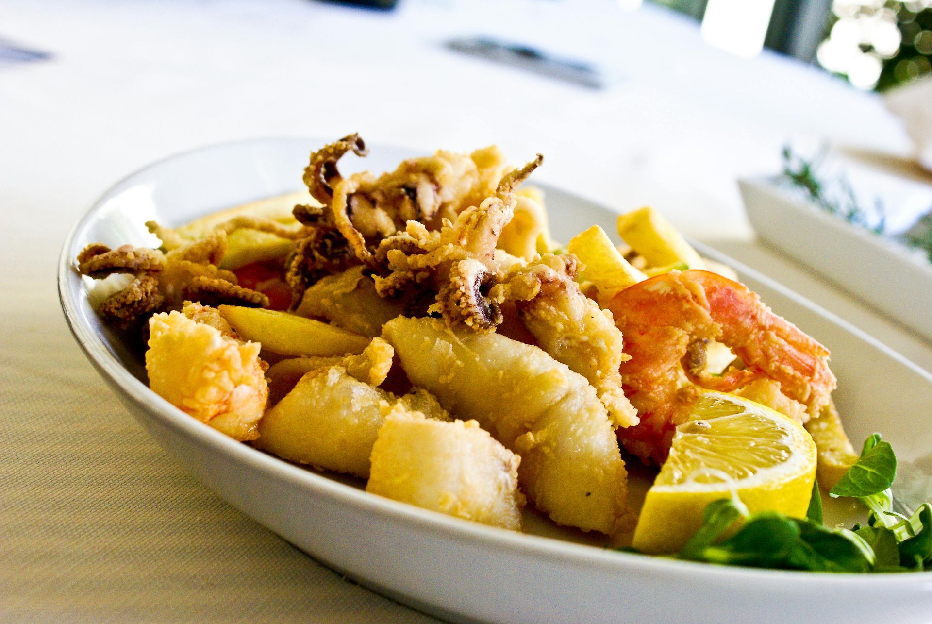 Piatto di frutti di mare con calamari fritti, gamberi, spicchi di limone e verdure su un piatto bianco.