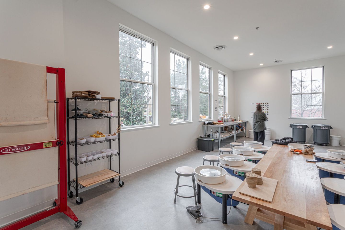 Bright pottery studio with windows, work tables, pottery wheels, shelves, and a person working.