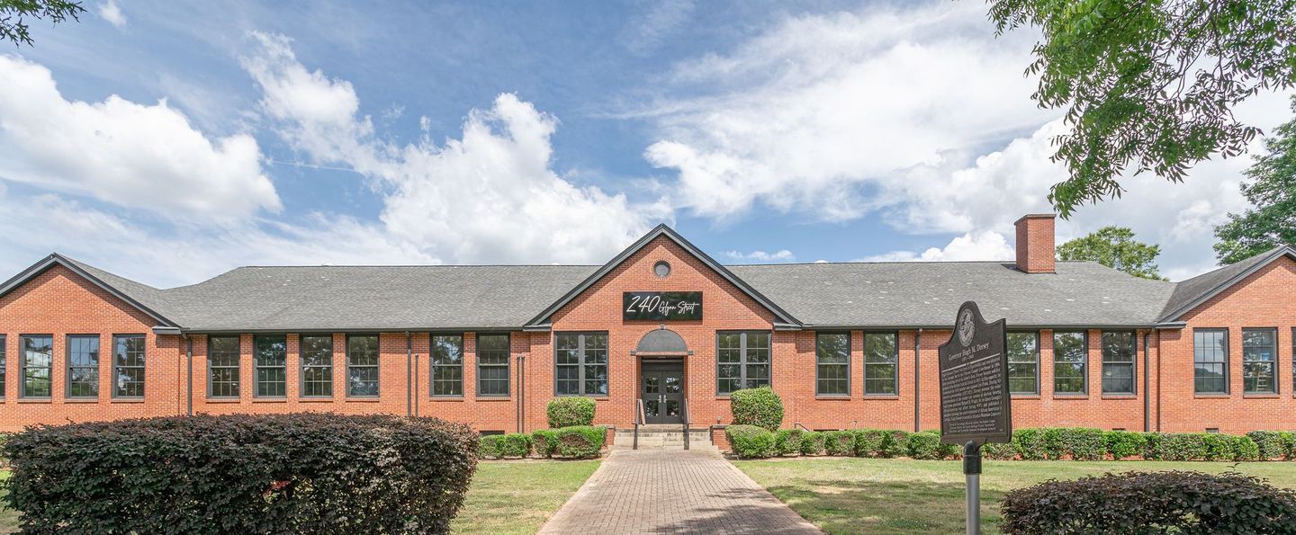 Red brick building with a central entrance and sign. Paved walkway leading to the doors. Green shrubs and blue sky.