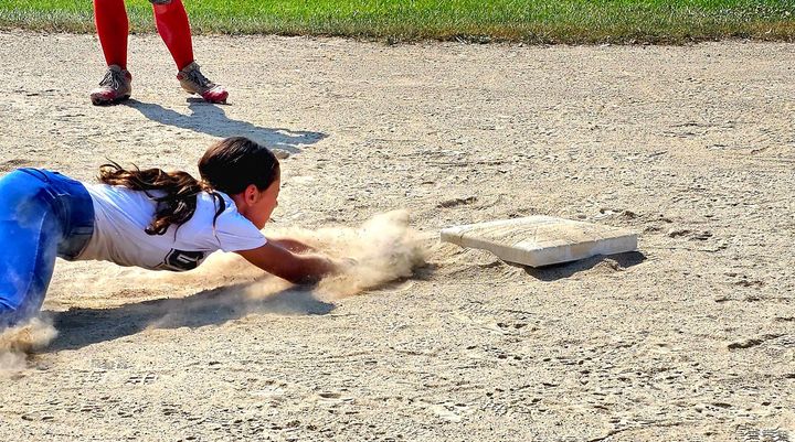 Girl sliding into a baseball base, kicking up dirt. Another player stands nearby.
