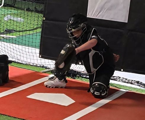 A young catcher in black gear kneels in the batter's box, ready to receive a pitch.