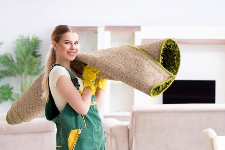 Woman carrying a rolled-up carpet in a bright living room, wearing green overalls and yellow gloves