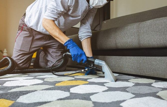 Person vacuuming a gray patterned rug beside a sofa with a handheld cleaner