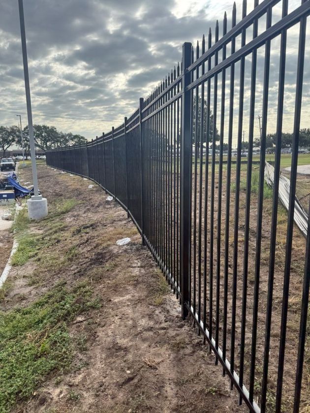 Black metal fence along a muddy roadside beside an open field under a cloudy sky