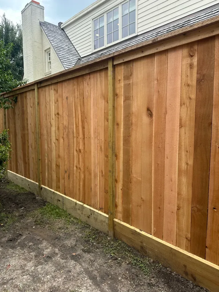 Wooden privacy fence beside a house, with new vertical planks and a dirt path along the base.