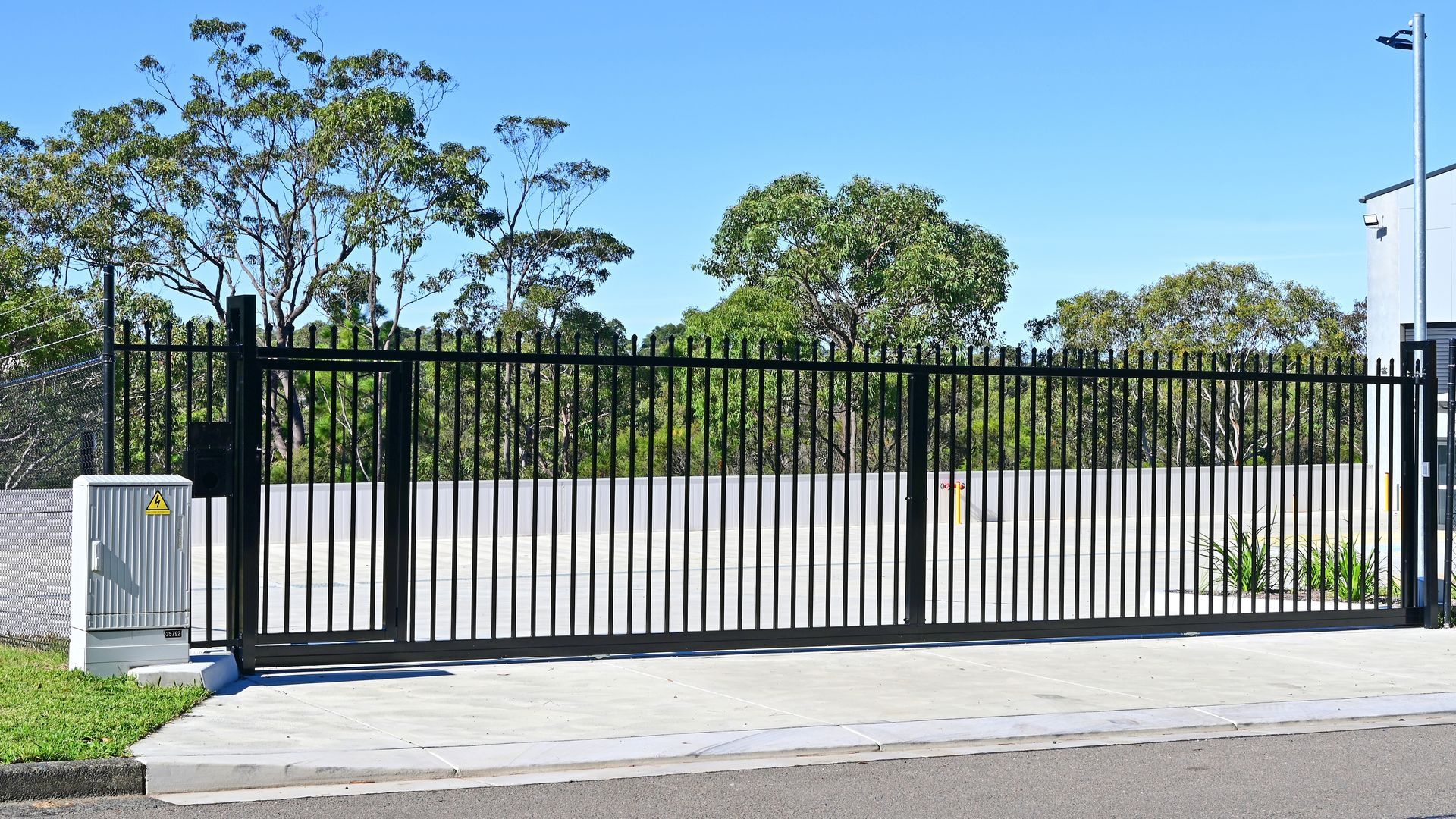 Black metal sliding gate along a concrete driveway, with trees and a road behind it.