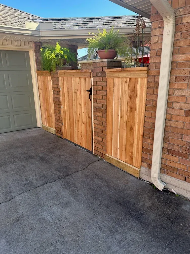 Wooden side gate and fence beside a driveway and garage, with a brick pillar and potted plant on top.