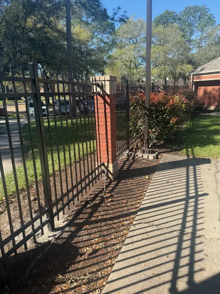 Black metal fence along a sunlit sidewalk beside a grassy yard and red brick building.