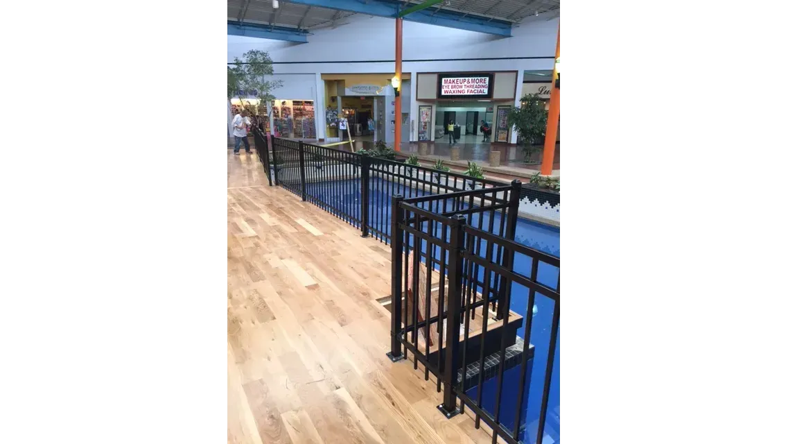 Indoor walkway with light wood flooring, black railings, and storefronts in a mall-like space