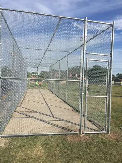 Chain-link baseball batting cage with concrete floor on a grassy field under a clear sky