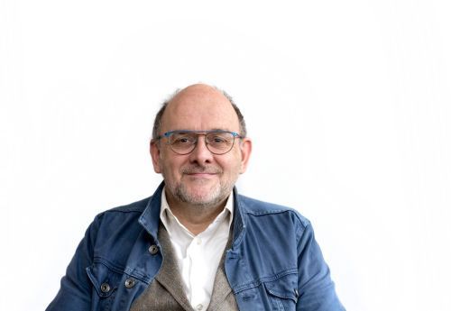 Portrait of a smiling person wearing glasses, a blue jacket, and a white shirt against a white background