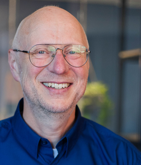 Smiling bald man in glasses and blue shirt, indoors with a blurred office background