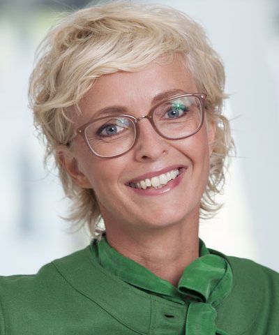 Smiling person with short blonde hair and glasses wearing a green jacket against a light background