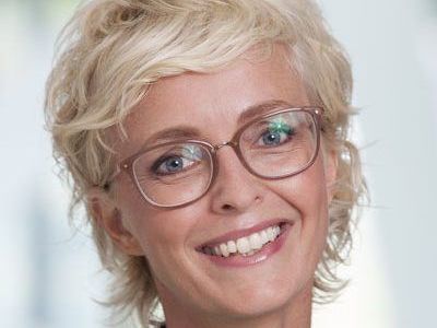 Smiling person with short blonde hair and glasses against a light background